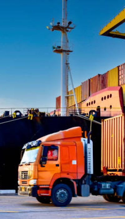 Truck carrying forty-foot container leaving port terminal with ship and quay crane on the background. Seaport operation activities, container shipping, and logistics.